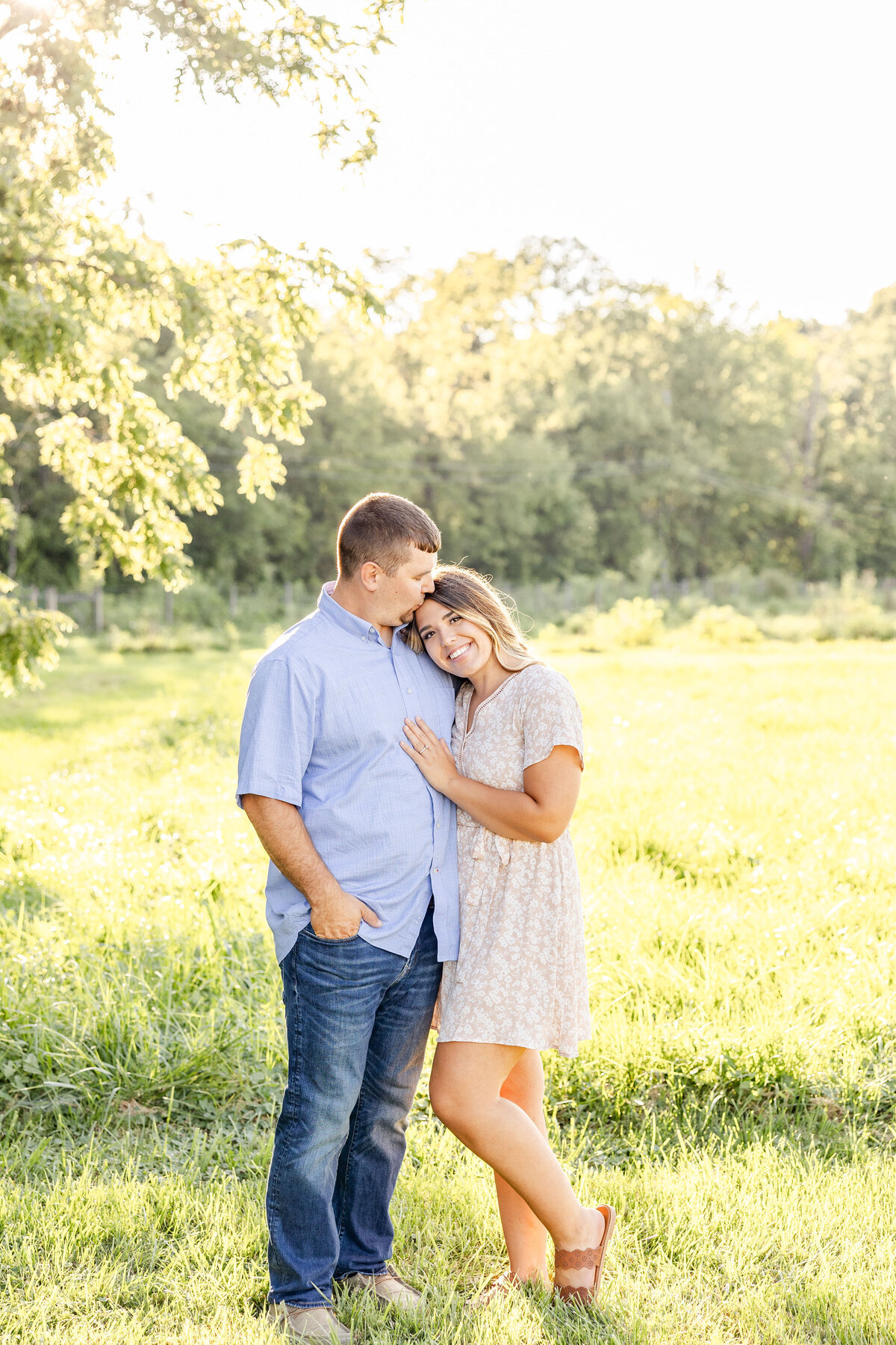Romantic husband and wife in a grassy field where he kisses her head with one hand in his pocket and one wrapped around her back and she smiles at the camera with one hand wrapped behind him and one hand on his chest.