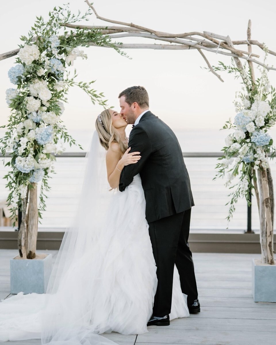 Wedding Photographer RI | A bride in a flowing white gown and a groom in a black suit share a kiss under a floral archway by the ocean.