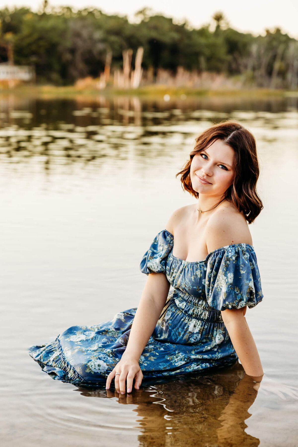 Gorgeous high schooler in a blue floral dress sitting in a calm lake as she glances over her shoulder for her senior photography session