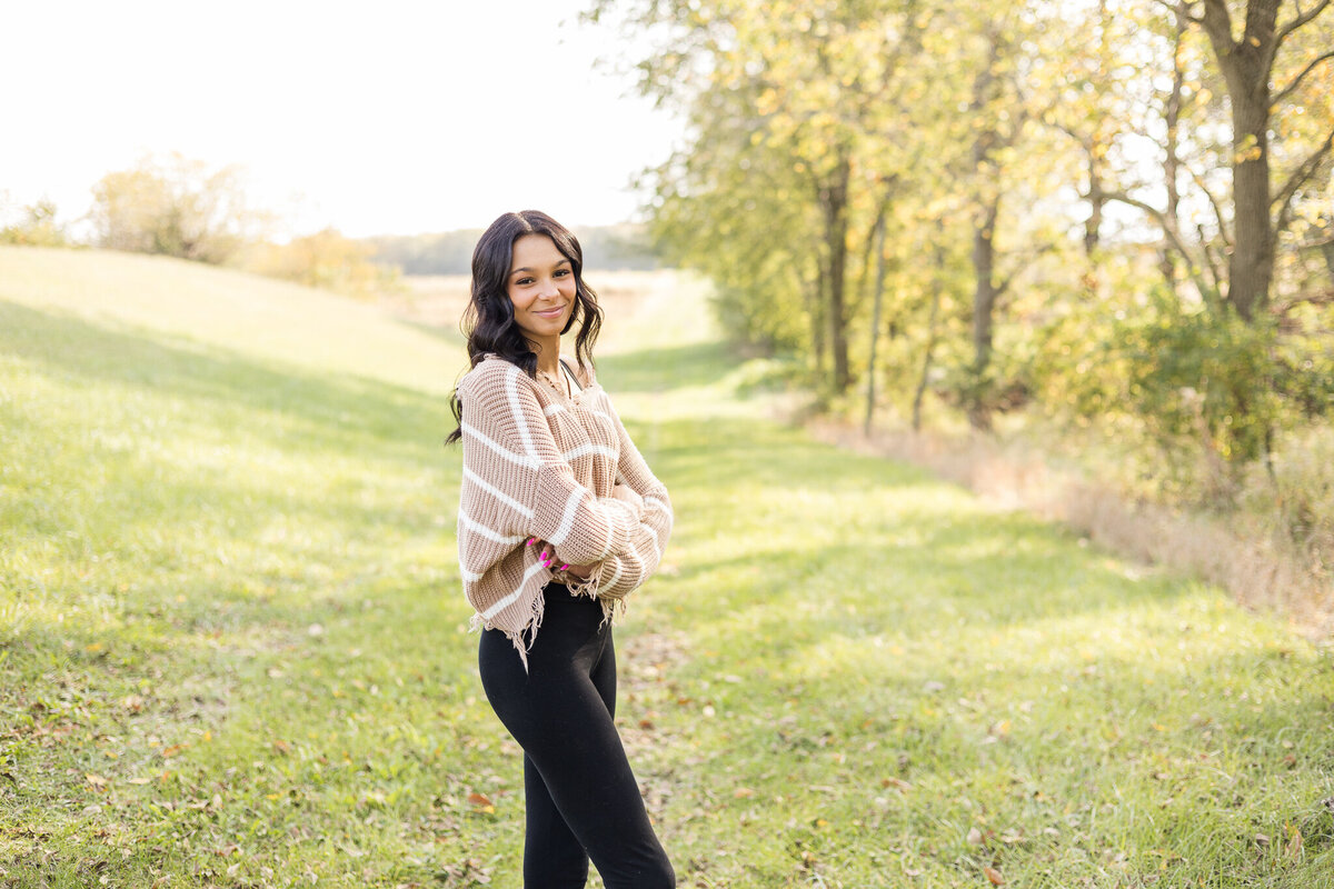 high school female senior, with long black hair, standing in grass with a tree line next to her, standing to the side, looking at the camera smiling softly, wearing black leggings, and light tan and white stripped shirt