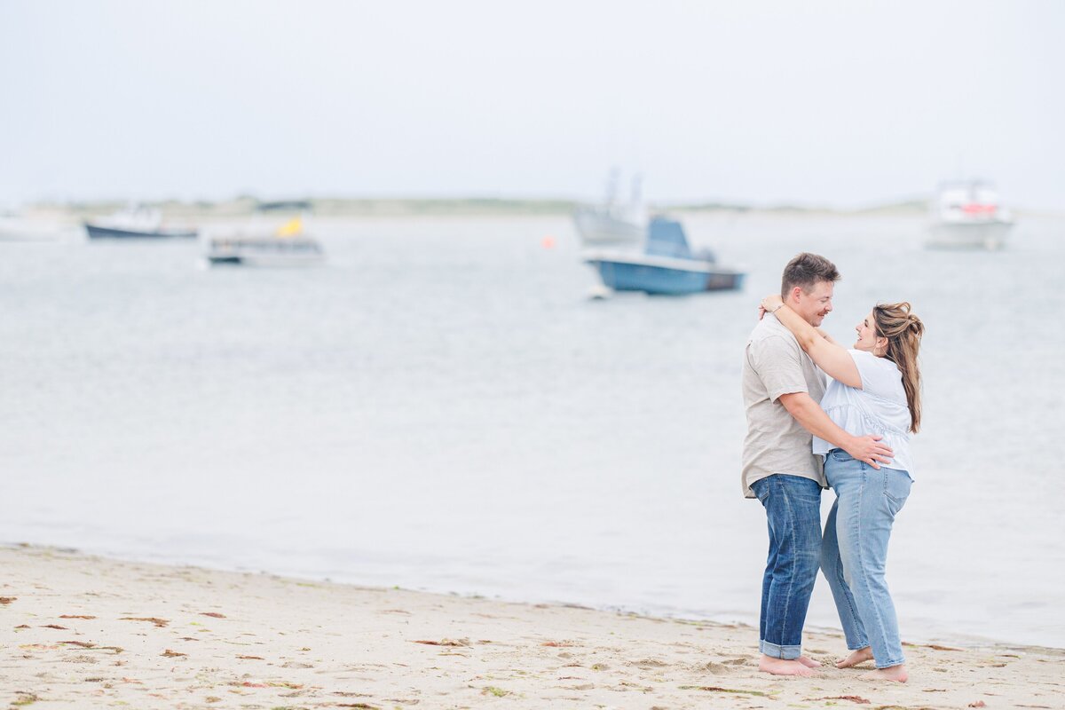 Couple standing barefoot on the beach, smiling at each other with their arms around each other’s waists, with boats anchored offshore in the background at Chatham Bars Inn in Chatham, Massachusetts — representing the portfolio of a Cape Cod engagement photographer.