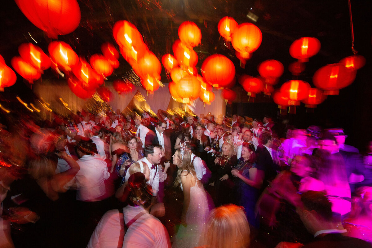 A lively wedding reception with guests dancing under a ceiling filled with glowing red lanterns.
