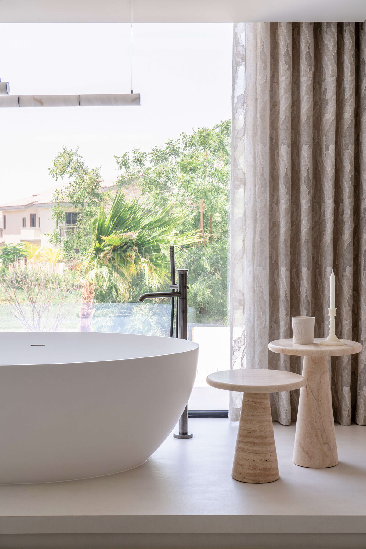 Large soaking tub in the primary bathroom. The wall behind the tub is entirely a window, looking out to a lush green scene. To the right of the tub is  two small, chic tables.