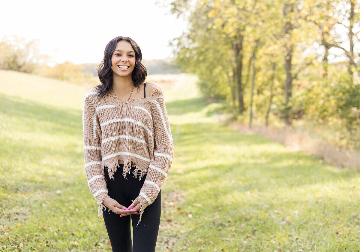 high school female senior, with long black hair, standing in grass with a tree line next to her, looking at the camera smiling, wearing black leggings, and light tan and white stripped shirt