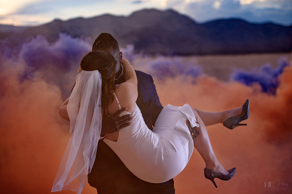 orange and  purple smoke bomb groom  carreis bride away as she wraps her arms around his neck las vegas elopement on the dry lake bed  at golden hour groom in blue suit jacket and black  pants  las vegas elopement eloping in vegas  las vegas wedding photographers las vegas wedding photography mk delacy photography