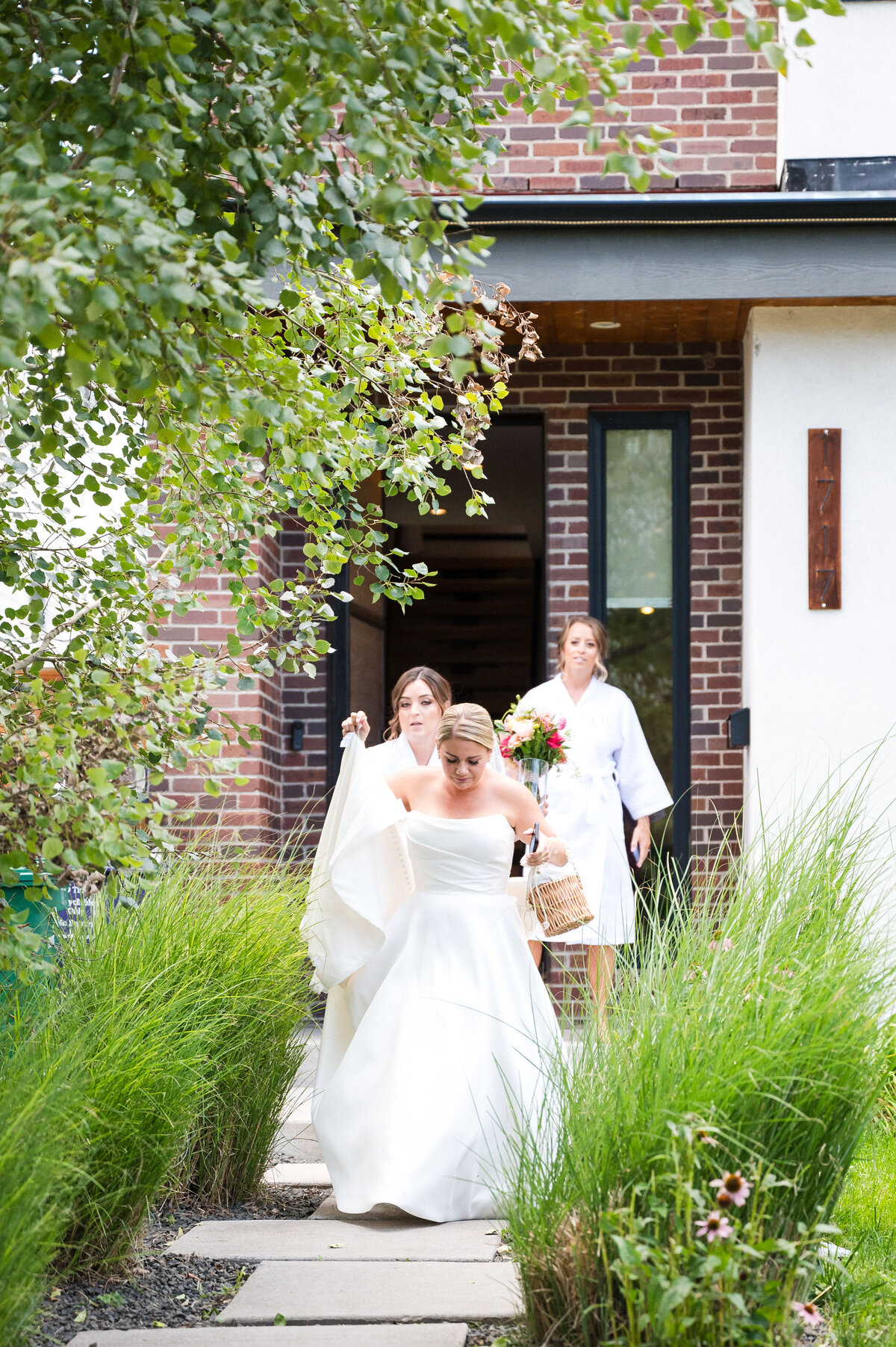 A bride exits her home in her wedding dress with two attendants helping with her dress.