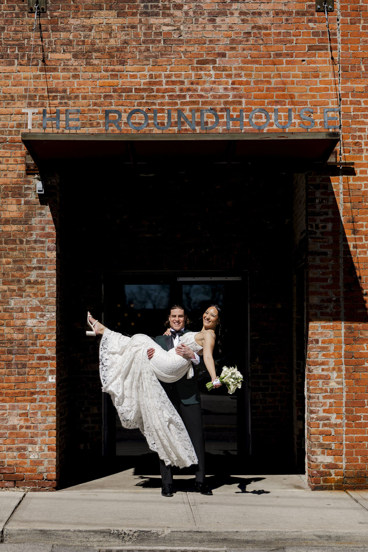 The groom picks up the bride and they are excited under the hotel sign at The Roundhouse