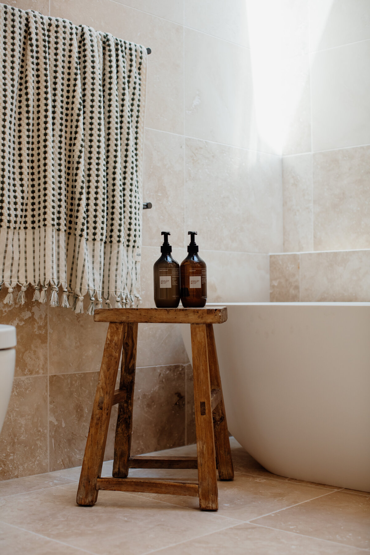 Bathroom designed by a Newcastle Interior Designer, featuring a wooden stool, two soap dispensers, and a towel on a rack. The white tub sits elegantly against tiled walls, showcasing luxury home design in Newcastle.