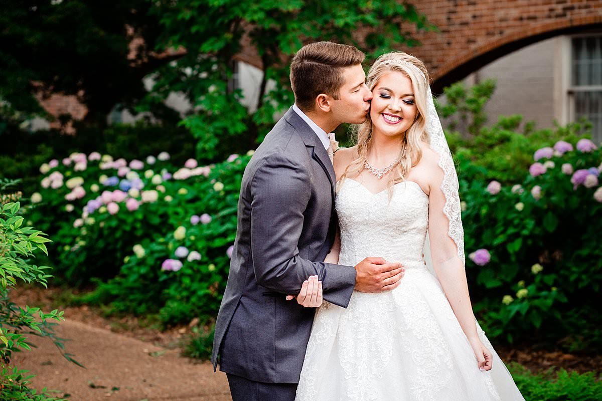 Groom kissing his wife on the cheek in the outdoor courtyard with hydrangeas behind them