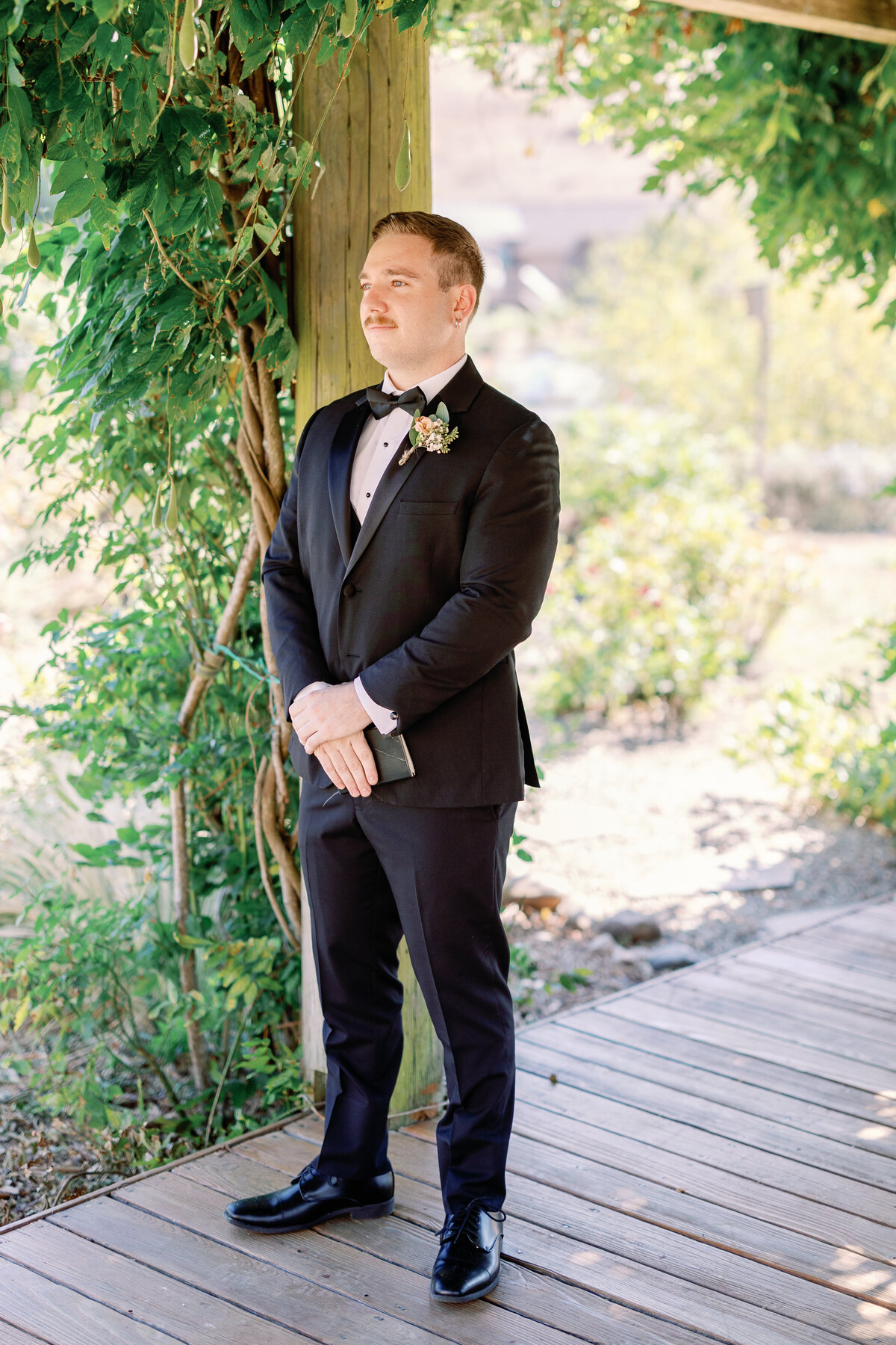 wedding photographer bay area captures groom standing in a black tux under a vine covered pergola waiting for his bride for their first look together