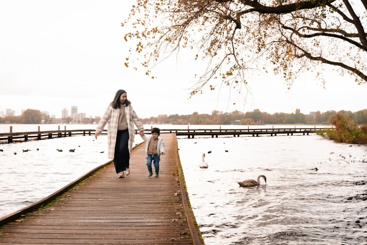 Mother holding hands with her young son while walking on a wooden pier beside swans and city skyline.