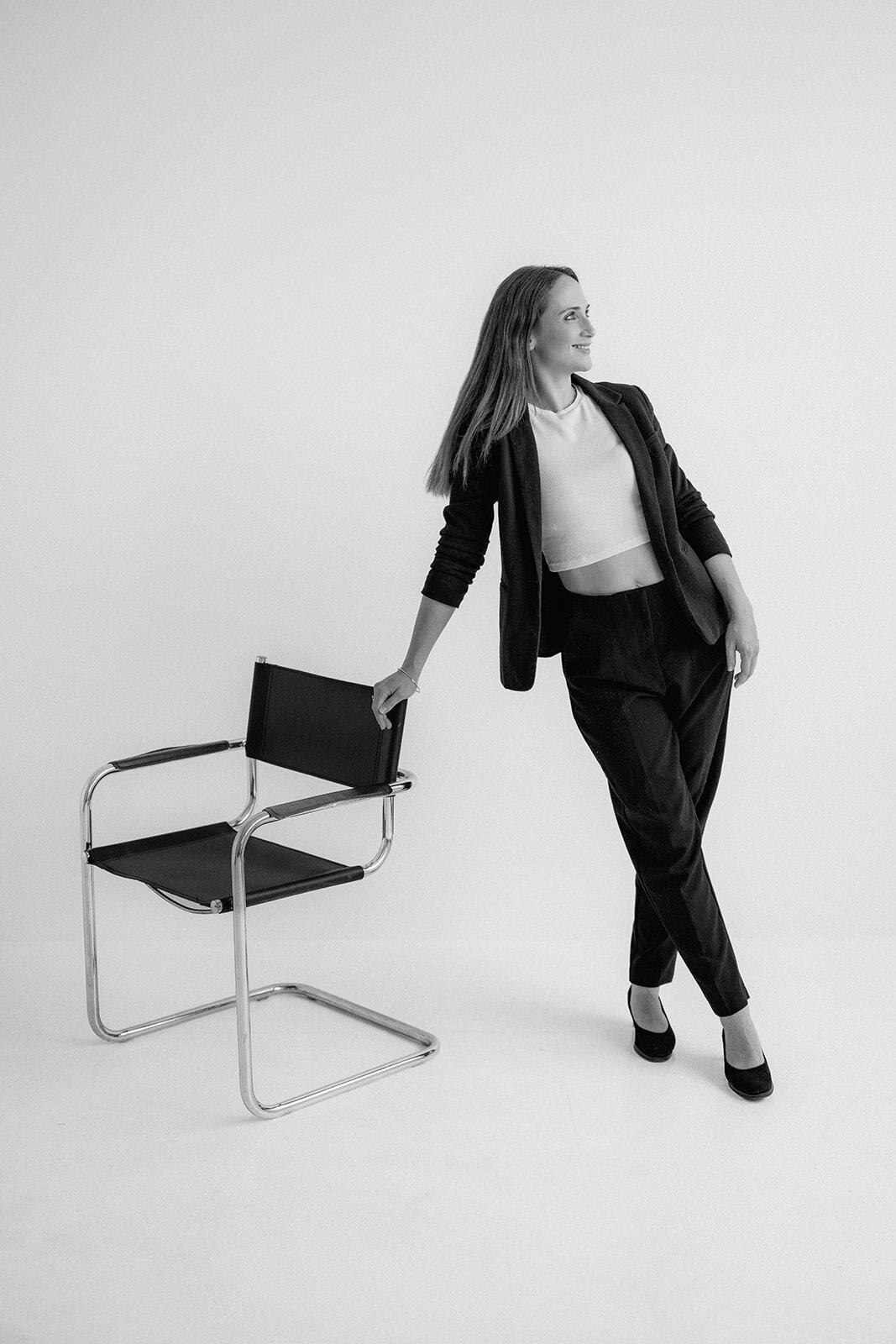 Woman standing next to a leather chair while leaning on it and gazing into the distance.