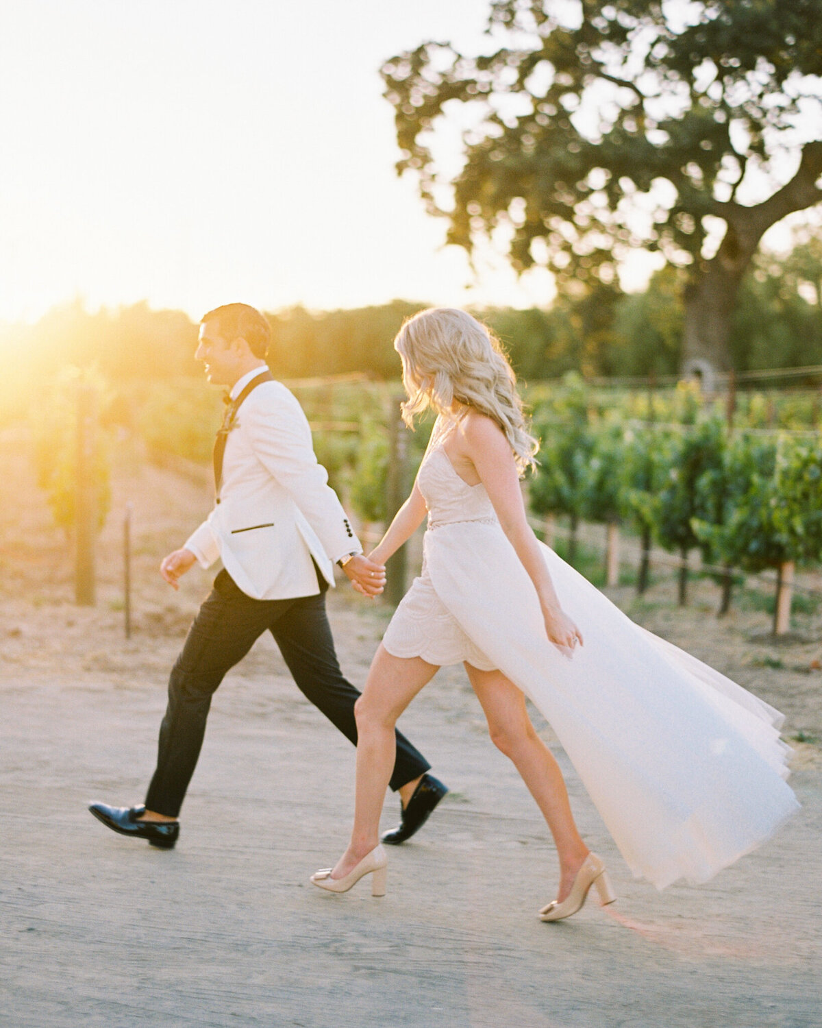 Trendy, modern bride and groom walking holding hands, stunning short wedding gown with detachable skirt, white suit jacket with black details, captured by Justine Milton Photography, fine art  wedding photographer & videographer in Calgary Alberta. Featured on the Bronte Bride Vendor Guide.