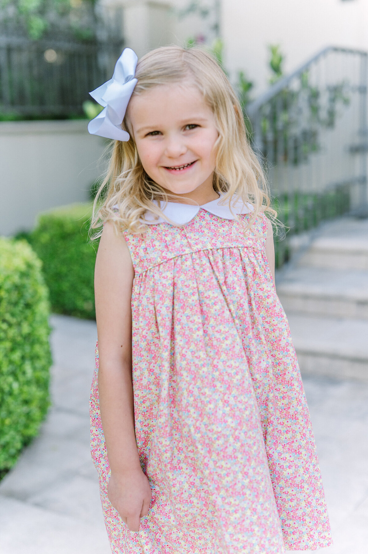 Five year old girl wearing a peter pan collared pink floral dress smiling in front of her home.