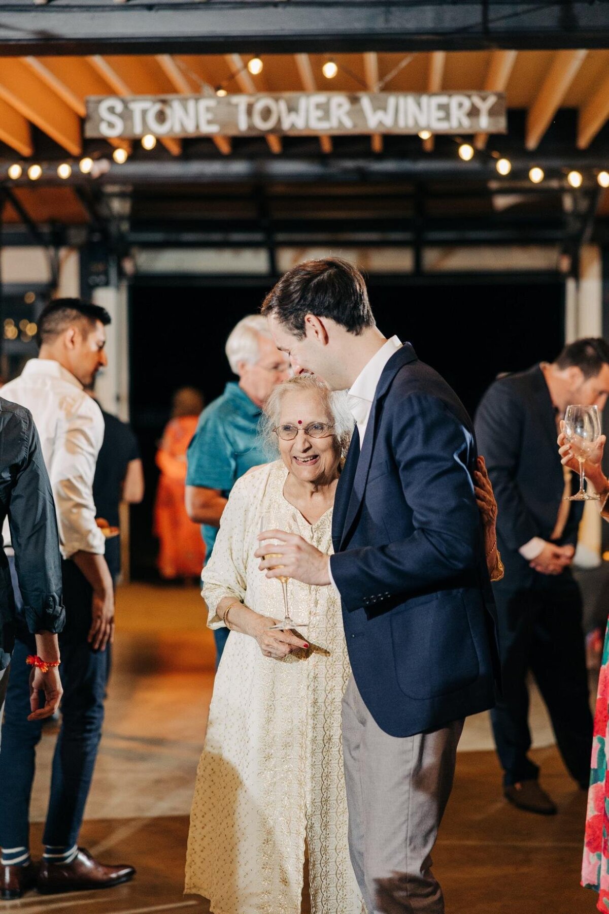 A young man gently hugs an elderly woman at a social gathering in a winery, both smiling warmly.