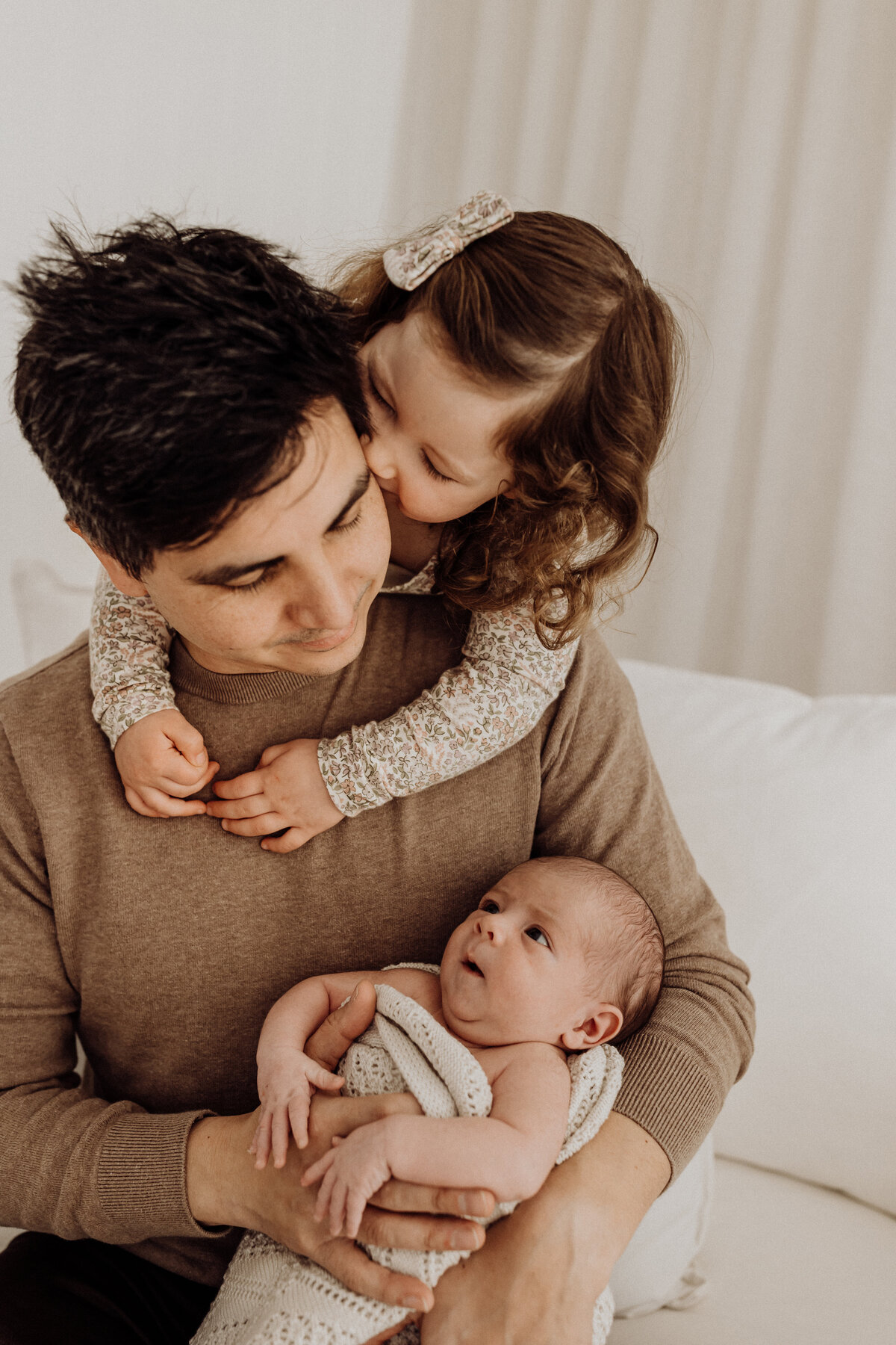 A father is hugged by his daughter while holding his newborn baby during a newborn photoshoot in a christchurch newborn photography studio