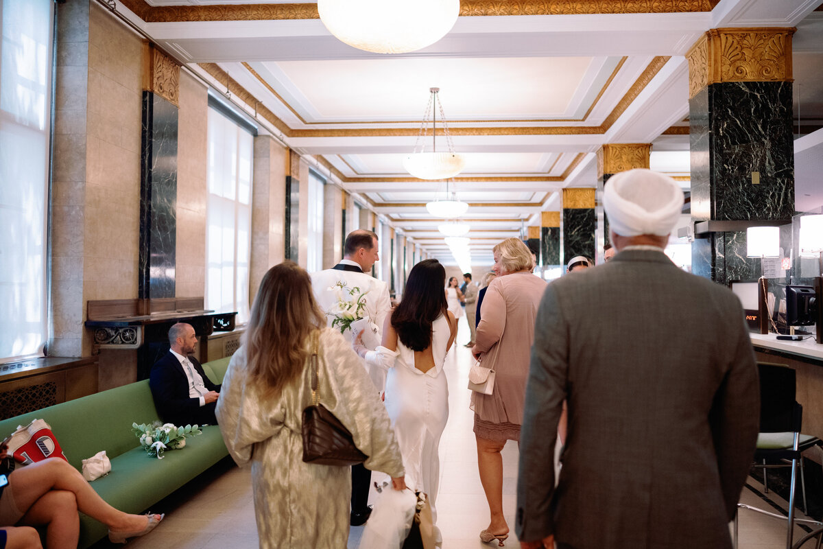 Bride and groom walking through the grand hallway of New York City Hall with family members during their elopement, captured candidly by NYC wedding photographer Perry Hancock.