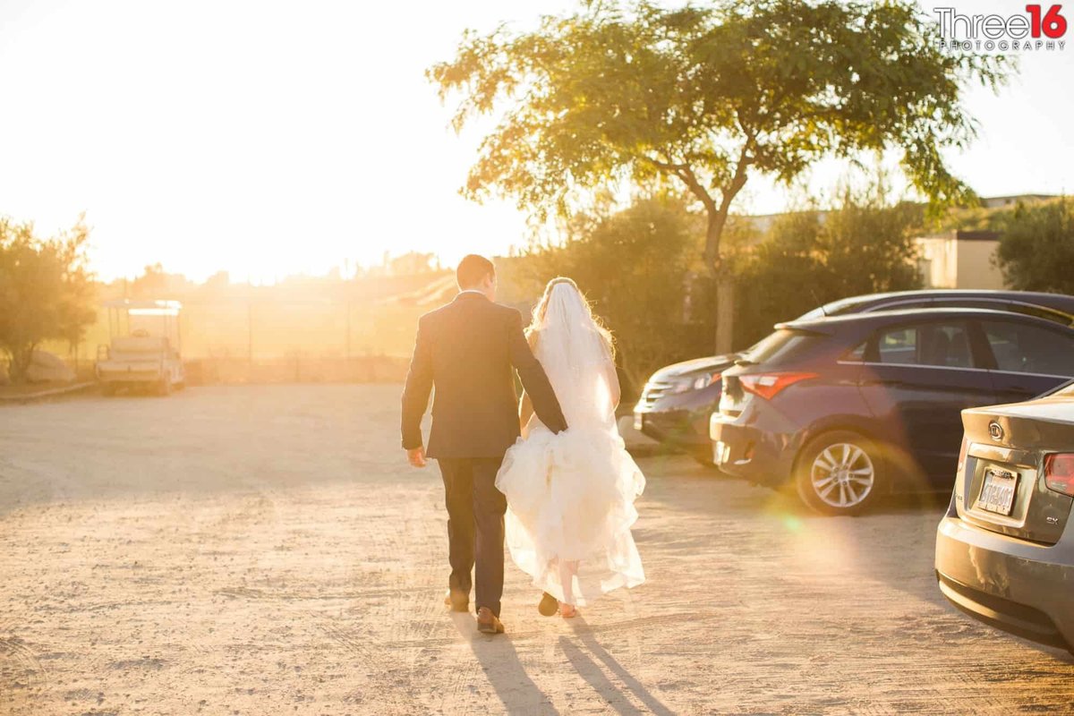 Groom carries his Bride's dress train across the dirt parking lot as they walk away