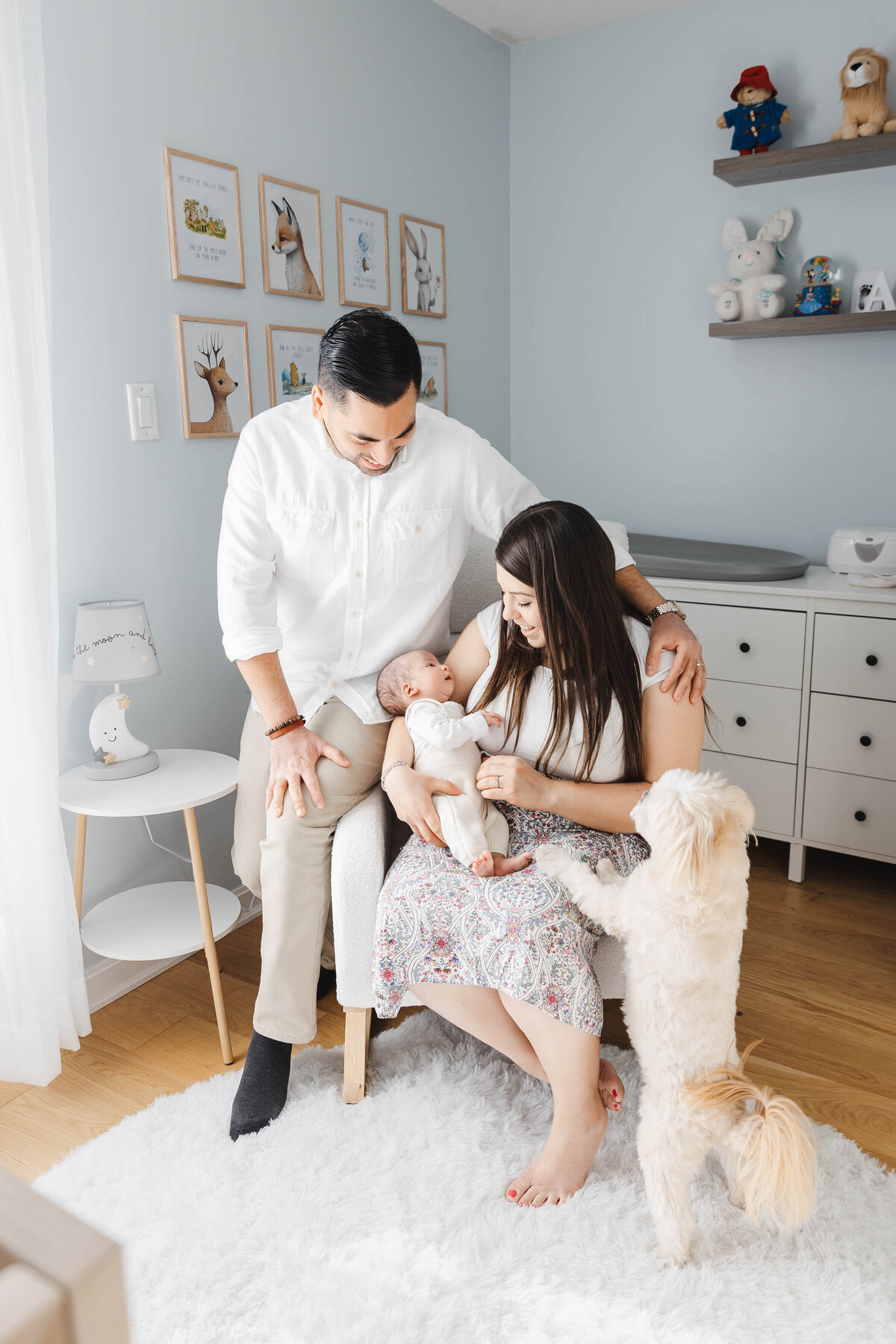 Mom holding newborn son in nursery rocking chair. Dad nestles on the armrest looking at his baby boy. Small white dog stands up to see the baby.