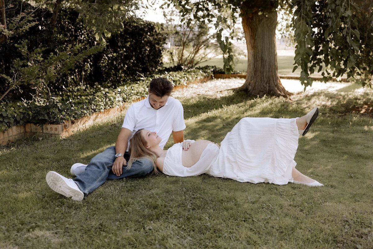 dad sitting in grass, pregnant mom laying in the grass with her head on his lap, hands on her exposed pregnant belly