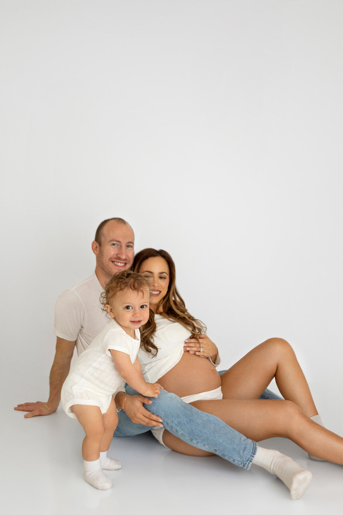 A joyful family portrait featuring a pregnant woman, her partner, and their toddler. The woman, dressed in a white crop top and blue jeans, sits on the floor with her bare baby bump visible. Her partner, wearing a white t-shirt and jeans, sits behind her, smiling warmly. The toddler, dressed in a white onesie and socks, stands in front of them, leaning on the woman's leg with a happy expression. The white background creates a clean and bright atmosphere, emphasizing the family's connection and anticipation of the new arrival.