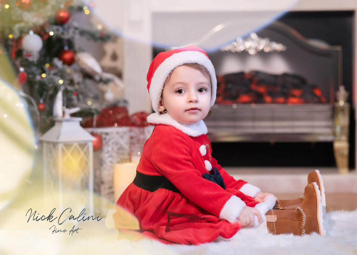 Baby dressed as Santa sitting by the fireplace in Calini Weddings Studio - festive Christmas setup with tree and warm lights.