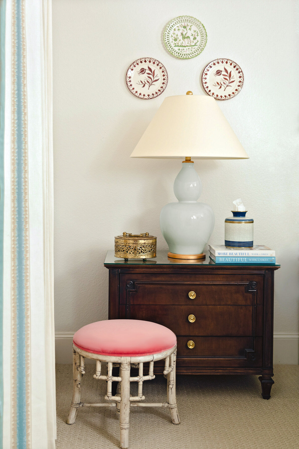 Wood nightstand with light blue table lamp and decorative accessories. Pink round stool with bamboo legs and decorative wall plates hung as wall decor.