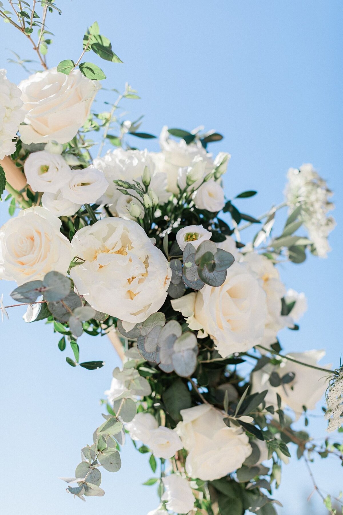 Close-up of a white rose and eucalyptus floral arrangement under a clear blue sky, styled for a romantic outdoor wedding ceremony