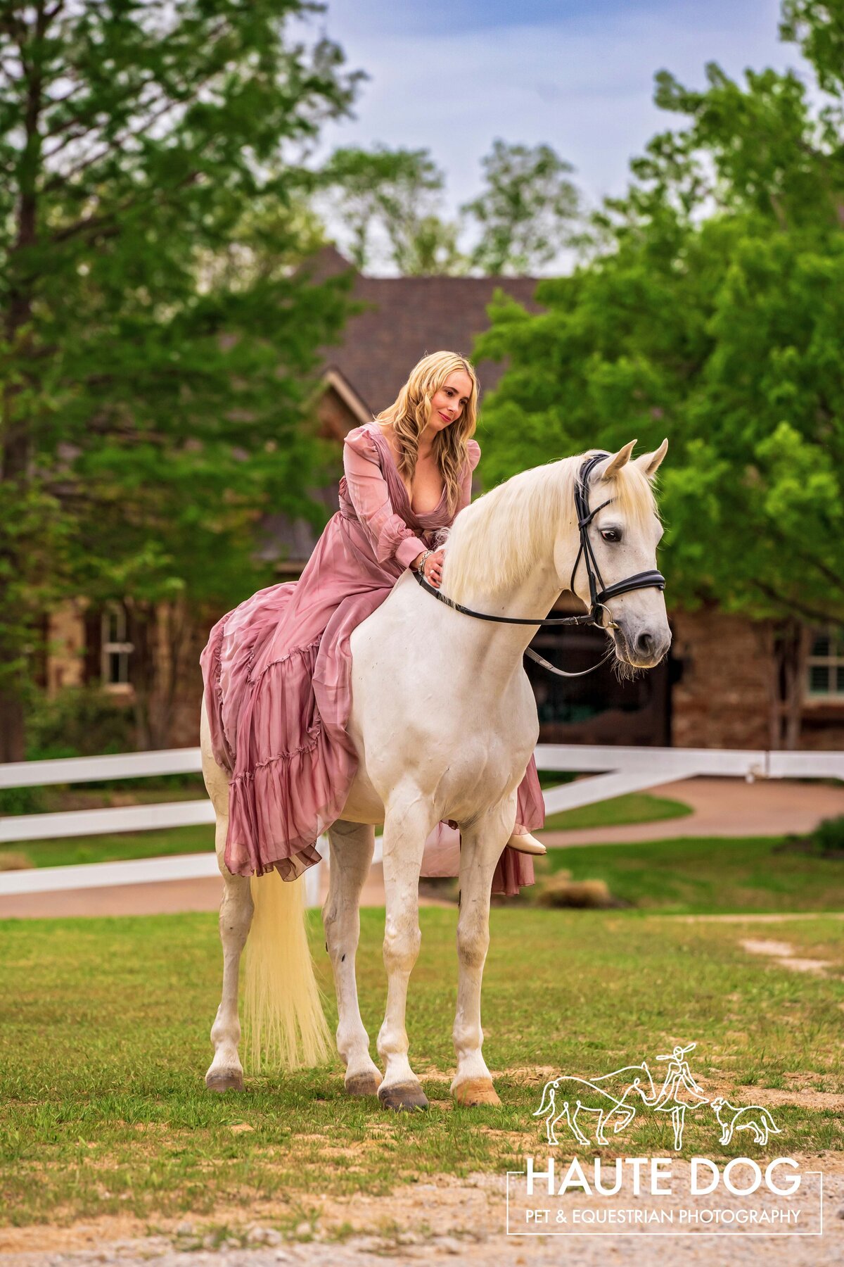 A woman wearing a long pink dress sits on her white gray horse and pats his neck.
