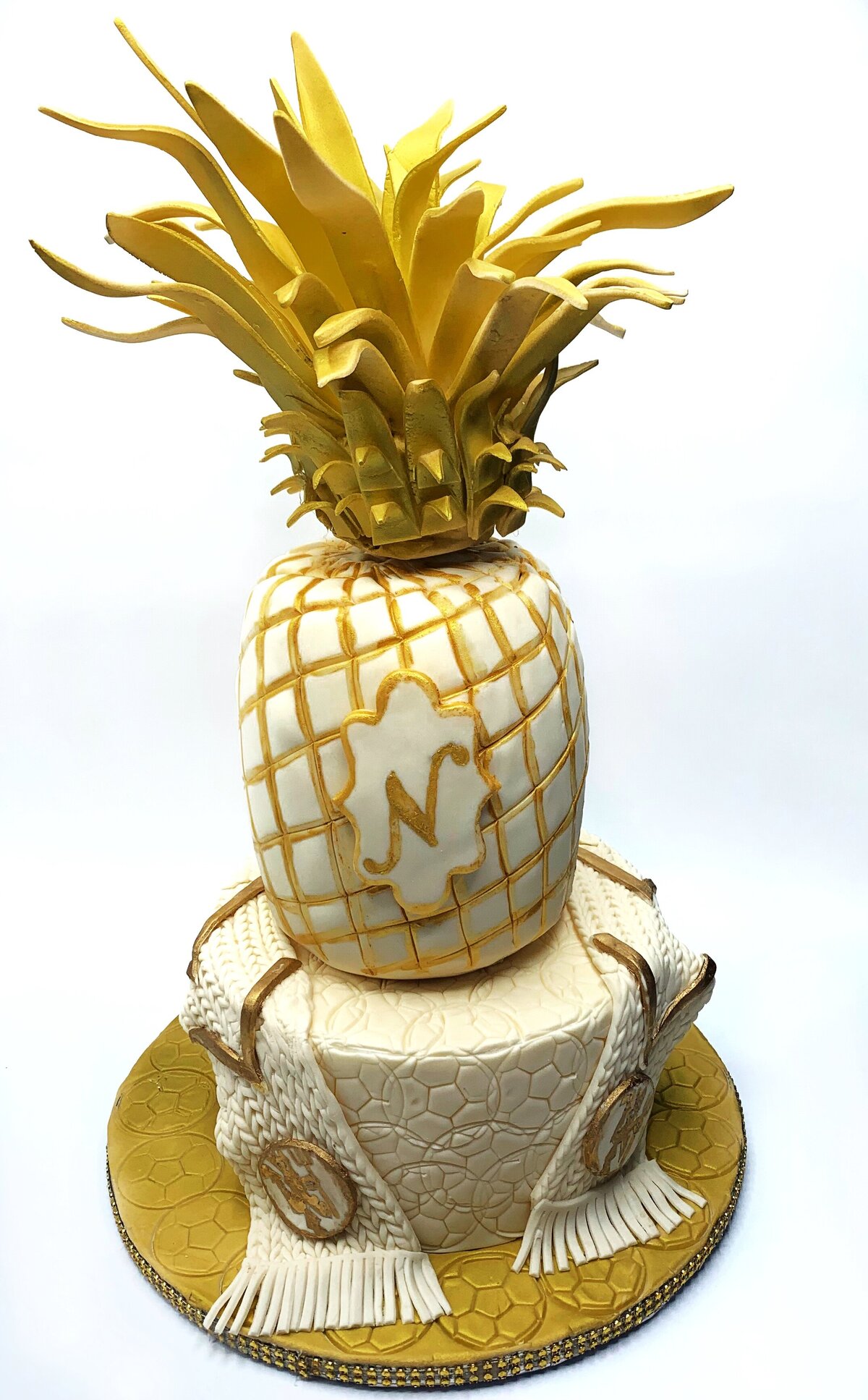 Whiet and gold 2 tier pineapple and soccer theme wedding cake. Top tier is white carved pineapple with gold accents and gold top leaves. Bottom tier is white with embossed soccer balls and wrapped in Manchester United soccer scarf with logos