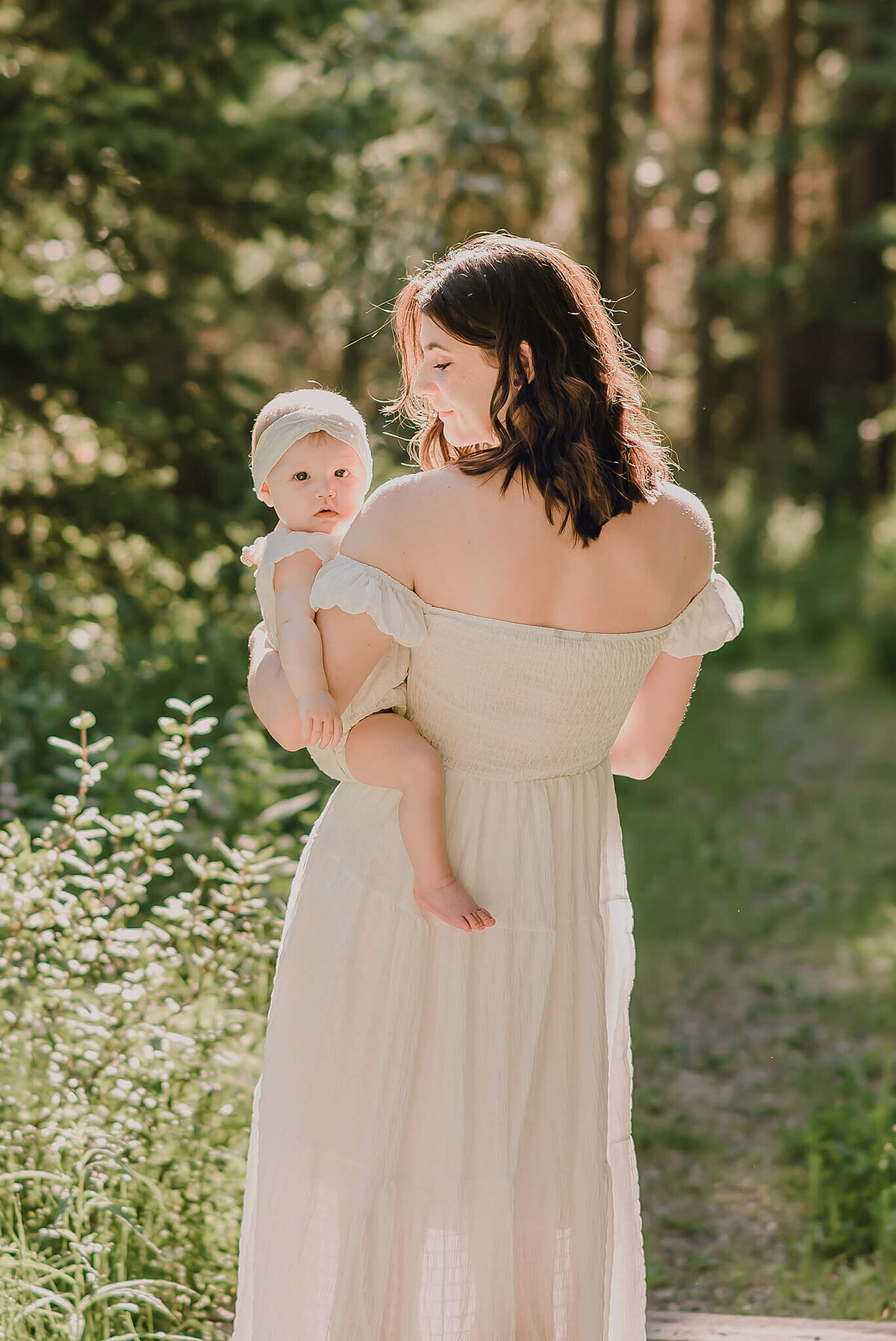 A beautiful image of a mother and baby daughter. Mothers back is to the camera while holding baby on her hip who faces the camera. They are backlit by beautiful sun. Captured by Brittany Danielle