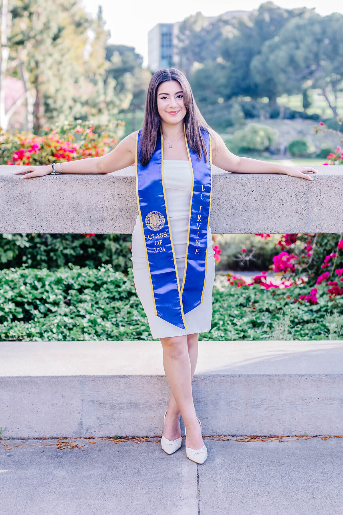 A woman wearing a blue graduation stole stands by a stone railing in front of a garden. She is smiling, with her arms resting on the railing. The garden is vibrant with red flowers and greenery, and a building is visible in the background.