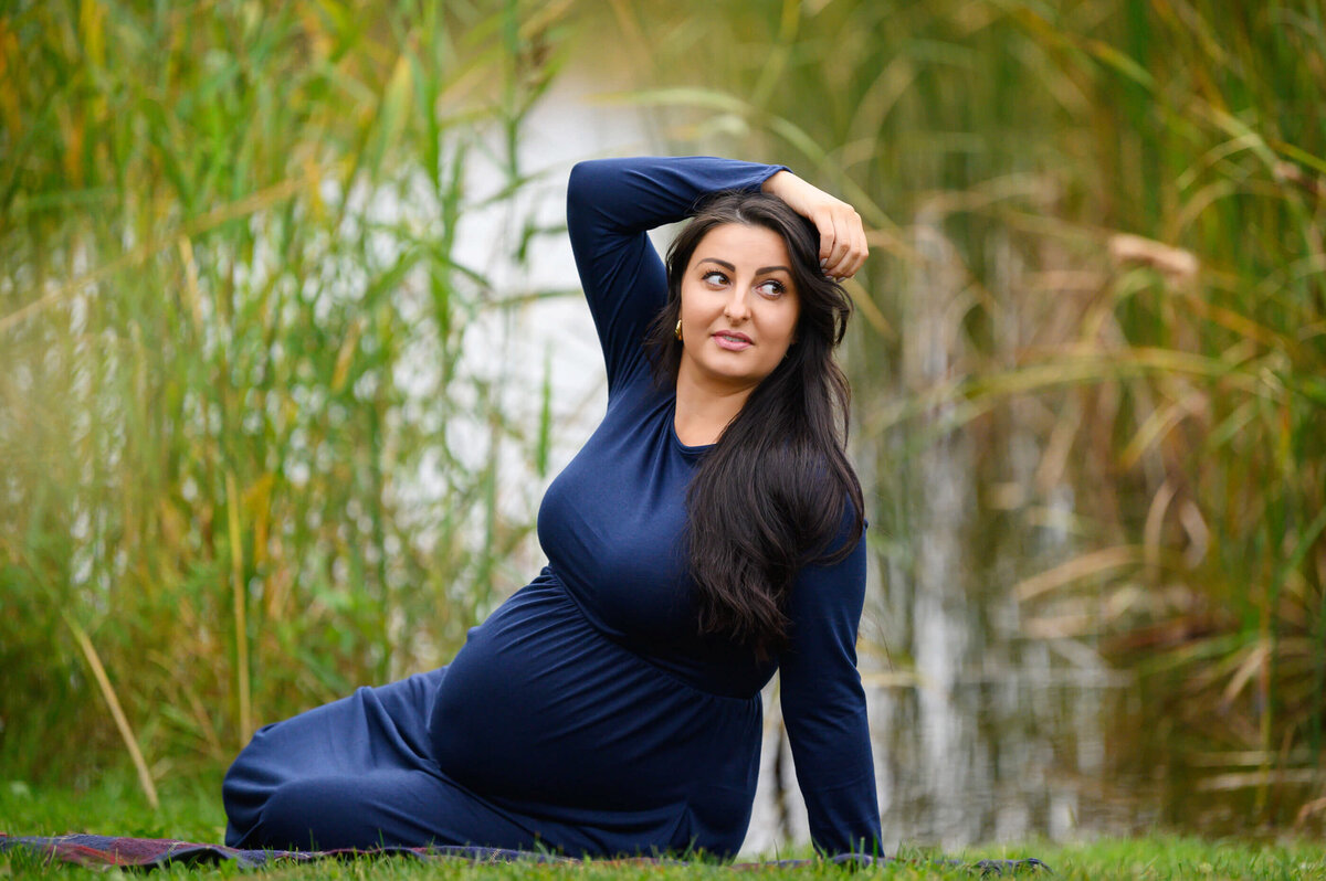 brown-haired woman sitting on the ground wearing a blue maternity dress for her fall outdoor maternity photography session in Burlington, On