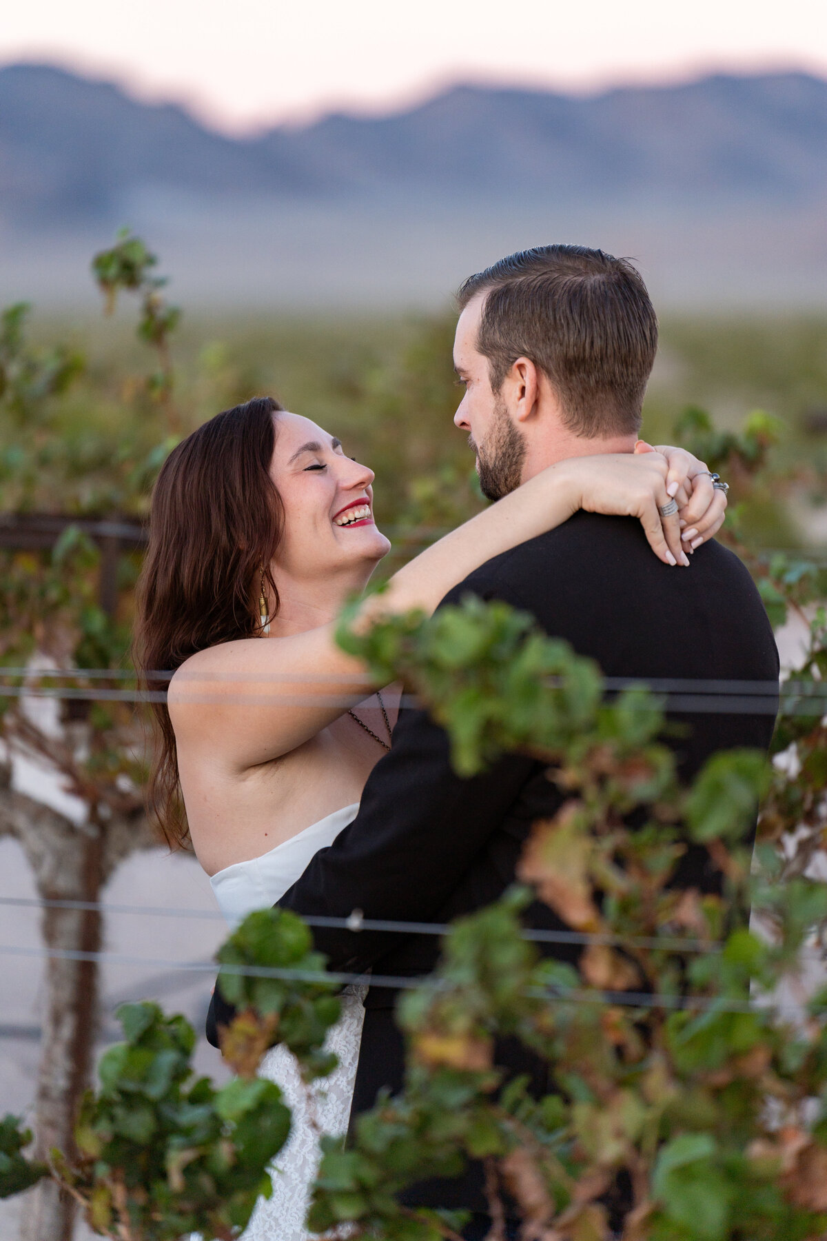 A couple standing in a vineyard smiling with their arms around each other