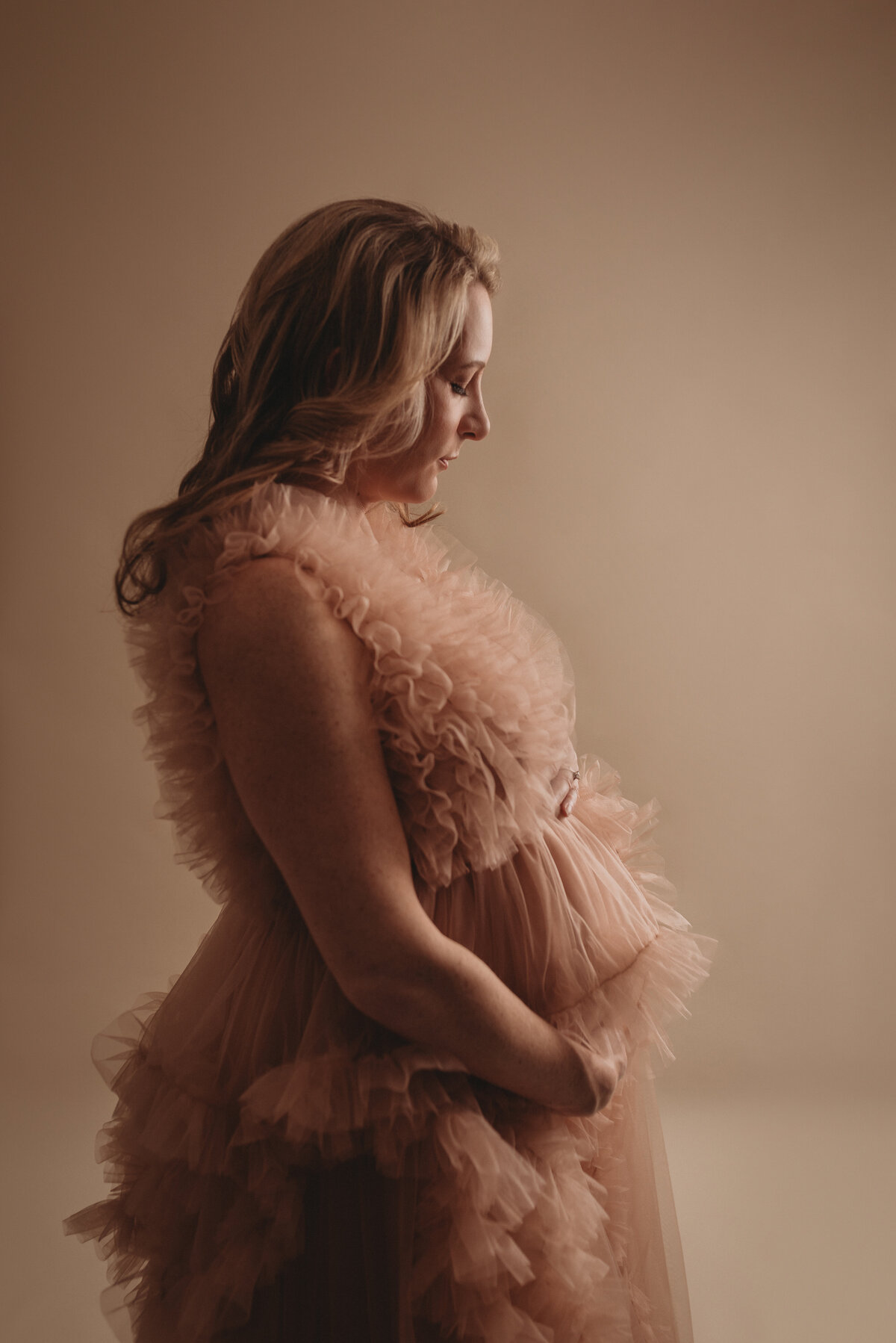 Blonde haired pregnant woman at Marietta GA maternity photography studio standing to the side showing tummy  in blush ruffle dress holding baby bump