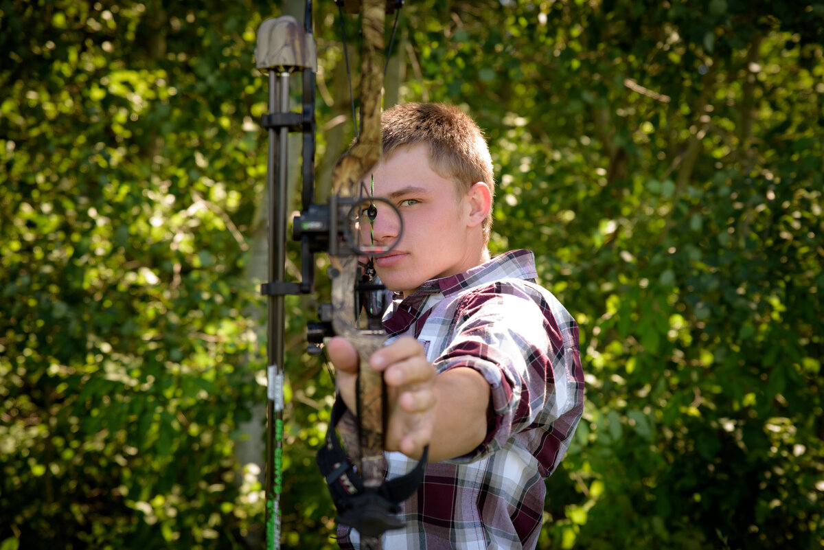 Luxemburg Casco high school senior boy wearing a maroon, black and white plaid button down shirt and jeans holding a hunting bow in farm field at his home near Green Bay, Wisconsin.