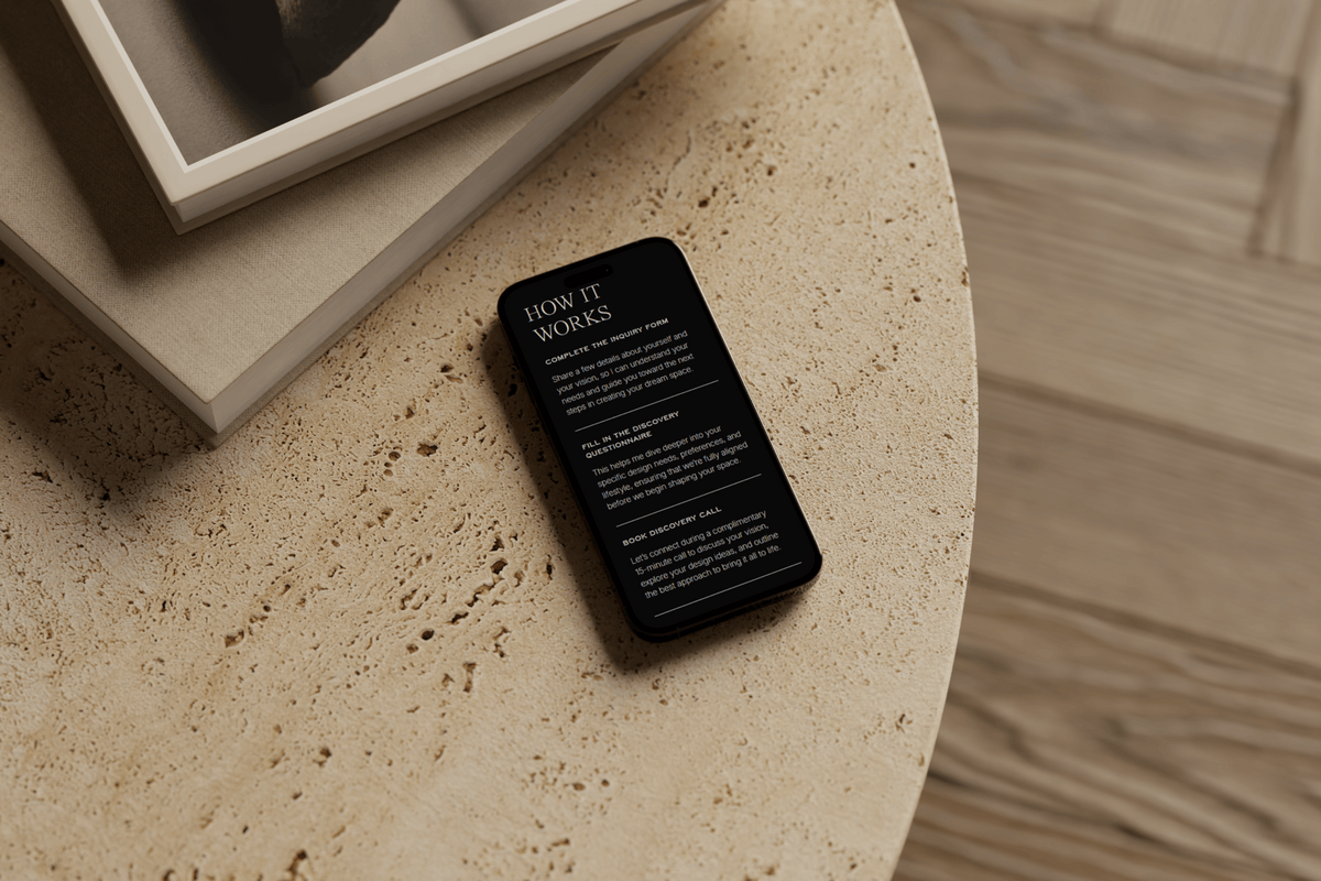 iPhone on a travertine coffee table, showcasing the "How It Works" details of virtual interior design services, with two stacked books beside it.
