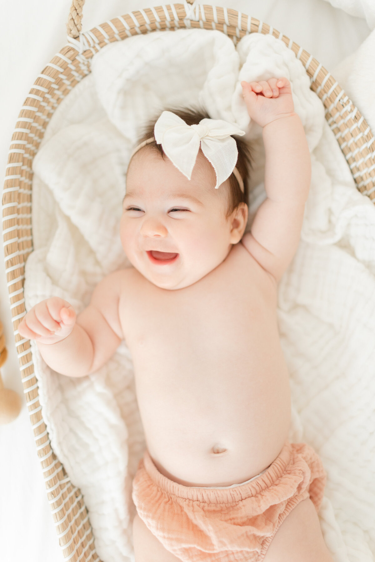 A Nova Studio Photography  photo of a sweet baby girl in peach bloomers with a white bow on her head laying in a basket smiling