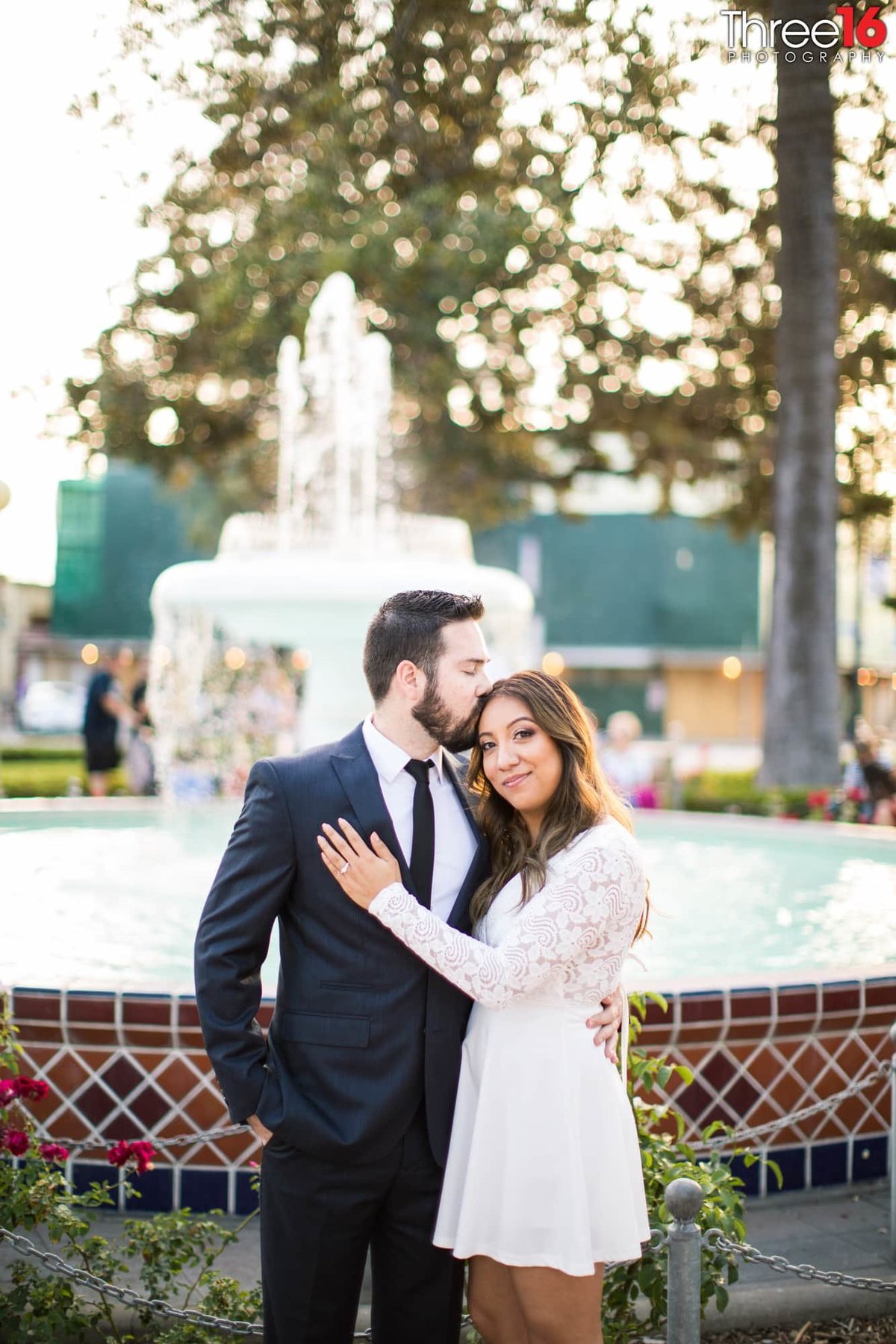 Groom to be kisses his fiance's head while standing in front of the Old Towne Orange Water Fountain