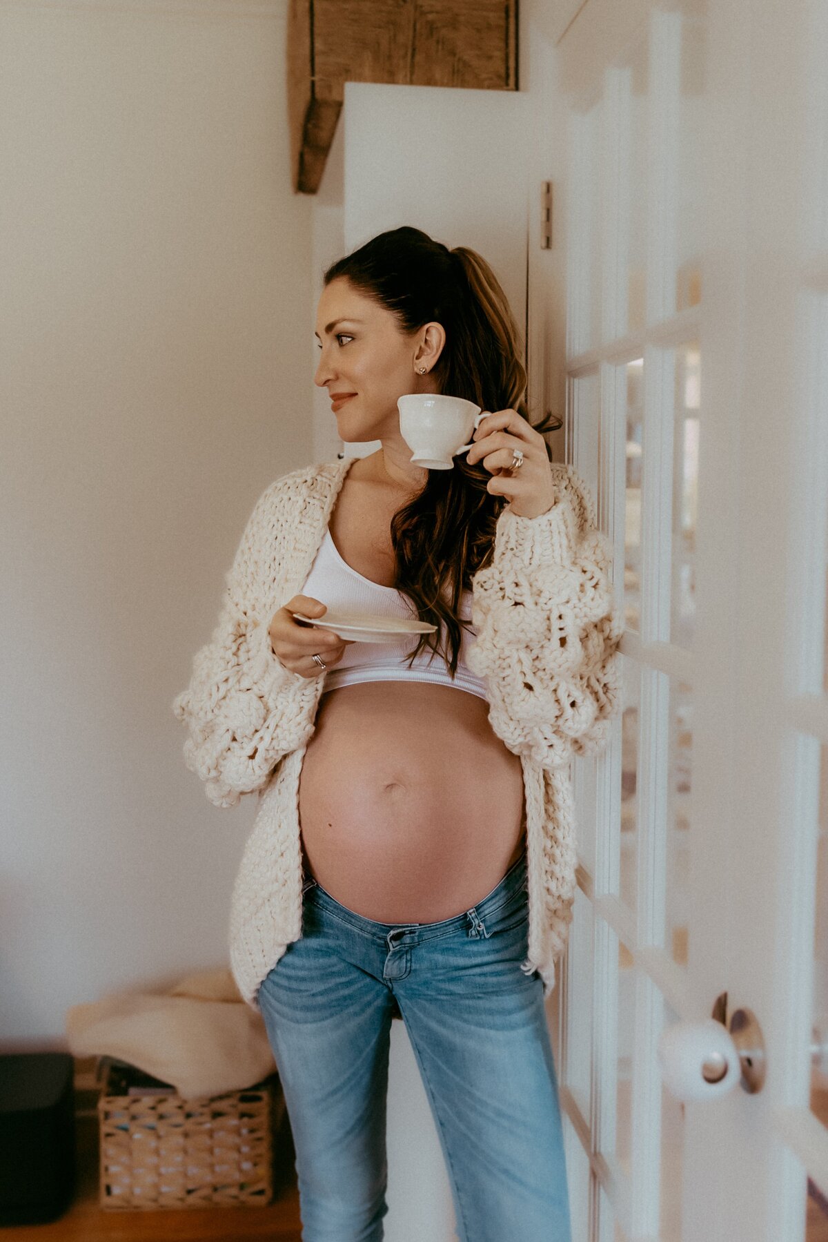 A woman with long hair, wearing a white top and jeans, stands by a door, holding a white cup and saucer. She is visibly pregnant and has a relaxed expression.