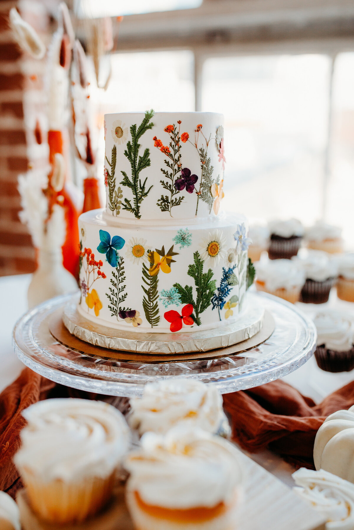 Colorful wildflower-themed wedding cake on display, surrounded by cupcakes on a dessert table