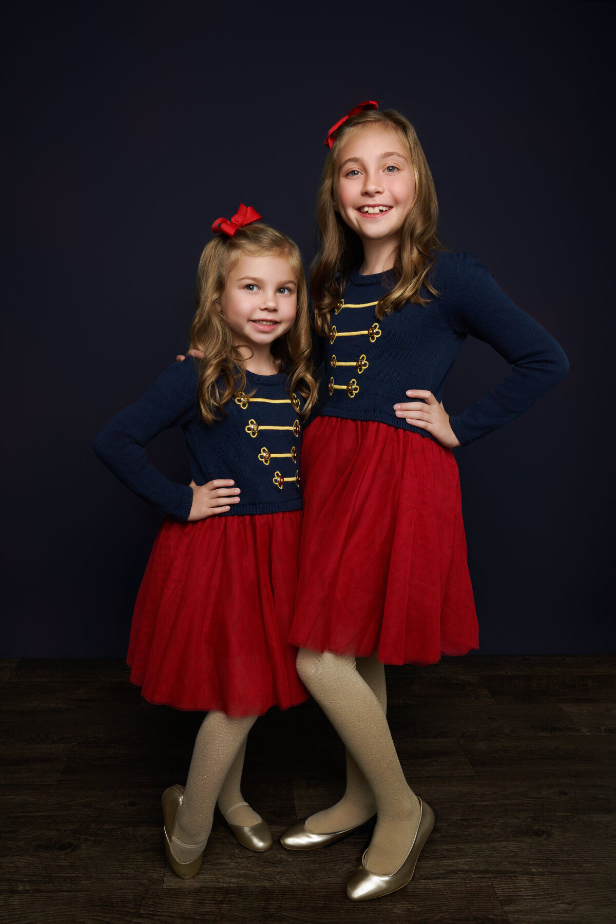 Sisters wearing Nutcracker inspired dresses posing in front of dark blue backdrop with Vanity Faire inspired lighting. 