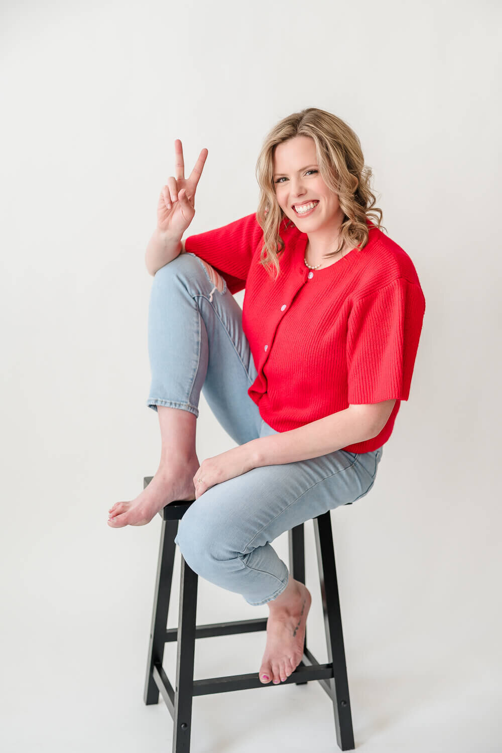 Smiling female entrepreneur giving peace sign while barefoot on stool, wearing red top and jeans.
