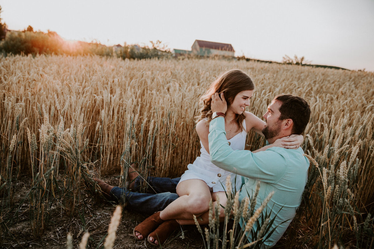 Woman sitting in man's lap in golden wheat at sunset. Her arms are wrapped around his neck and he is pushing the hair out of her face. There is a rustic Exeter, ON barn in the background.
