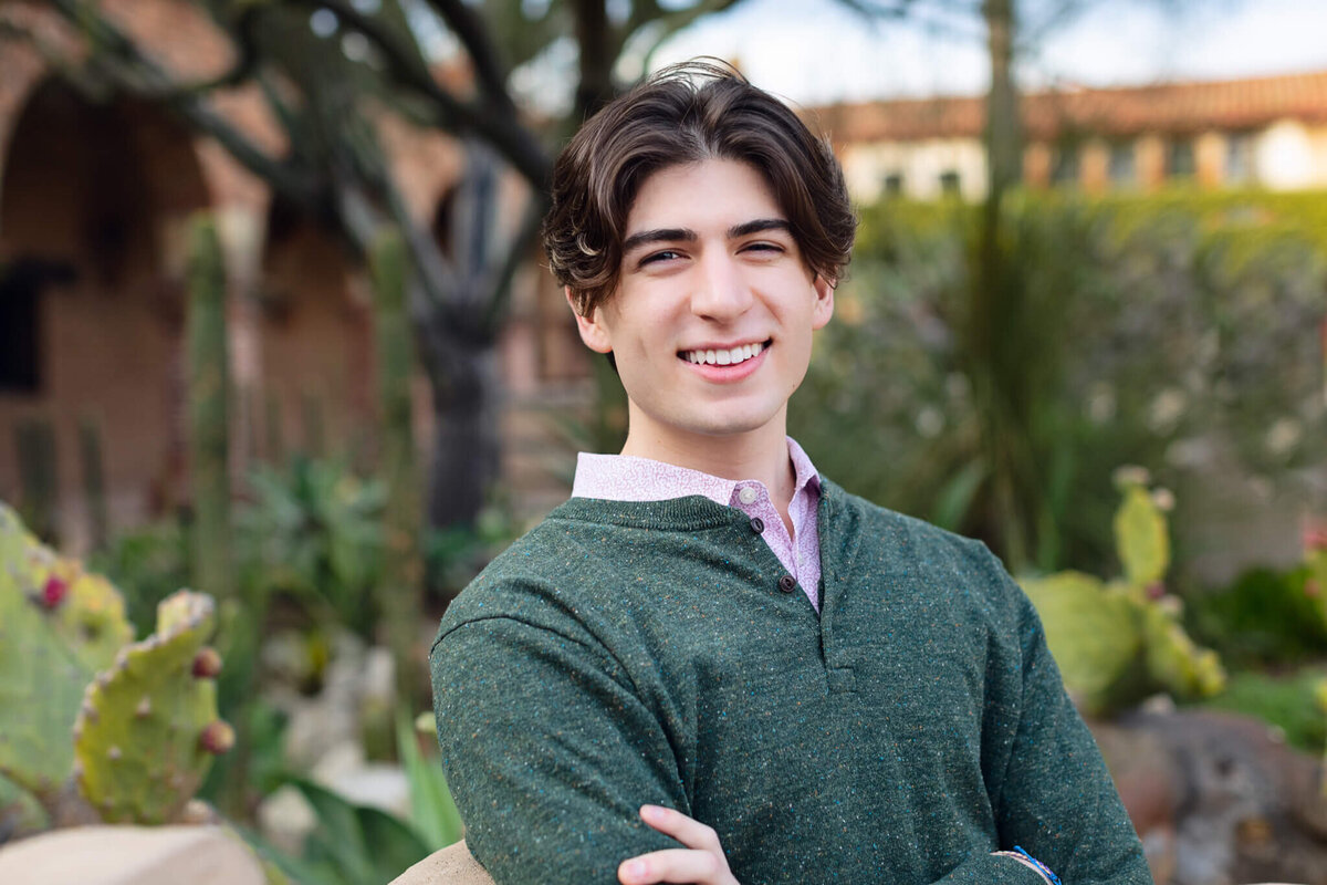 A young man wearing a green, buttoned shirt with dark hair smiling at the camera in a courtyard with greenery and brick walls.