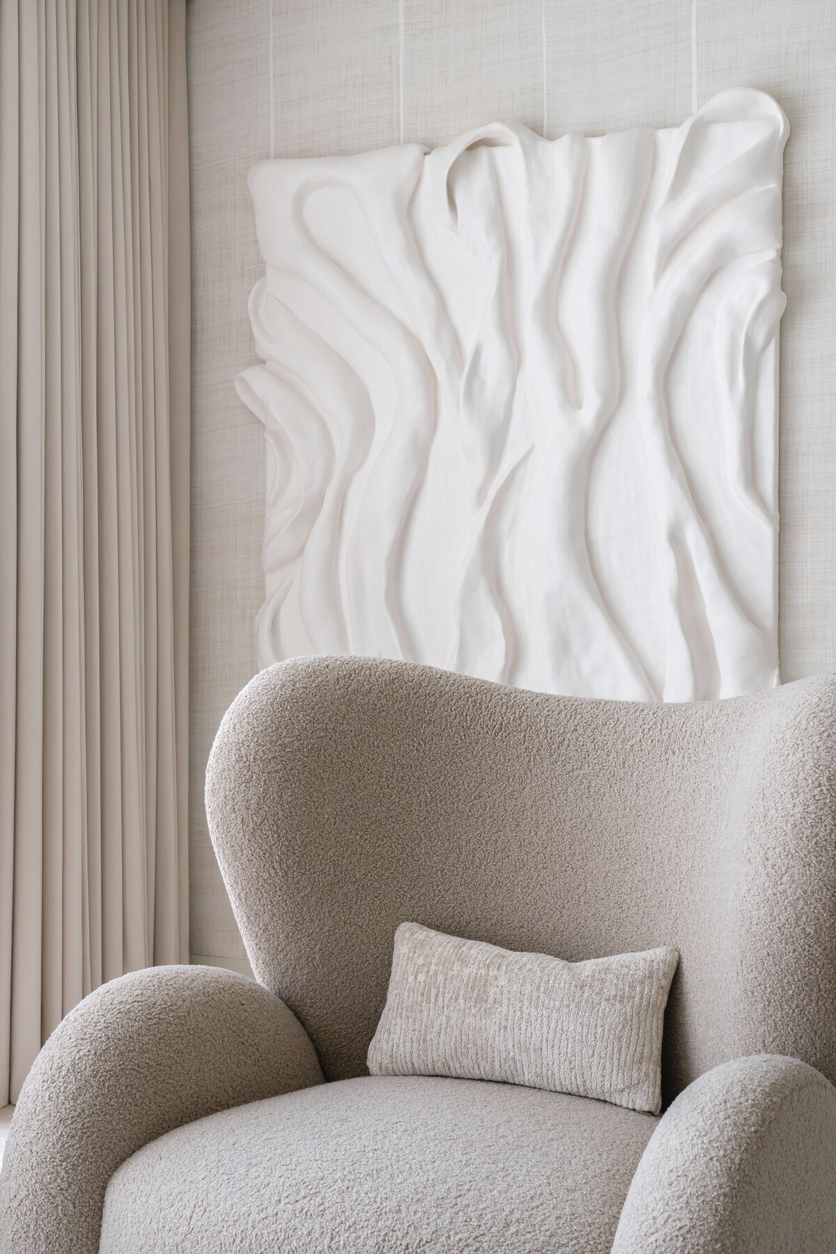 A chic neutral armchair in the primary bedroom. Behind the chair is a large contemporary white textured art piece.