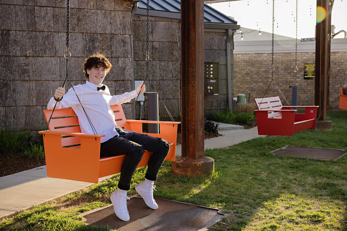 High school senior seated in a large orange adult bench swing wearing a white button down shirt, black jeans, white sneakers, and a black bowtie, captured with soft backlight by Charlotte photographer.