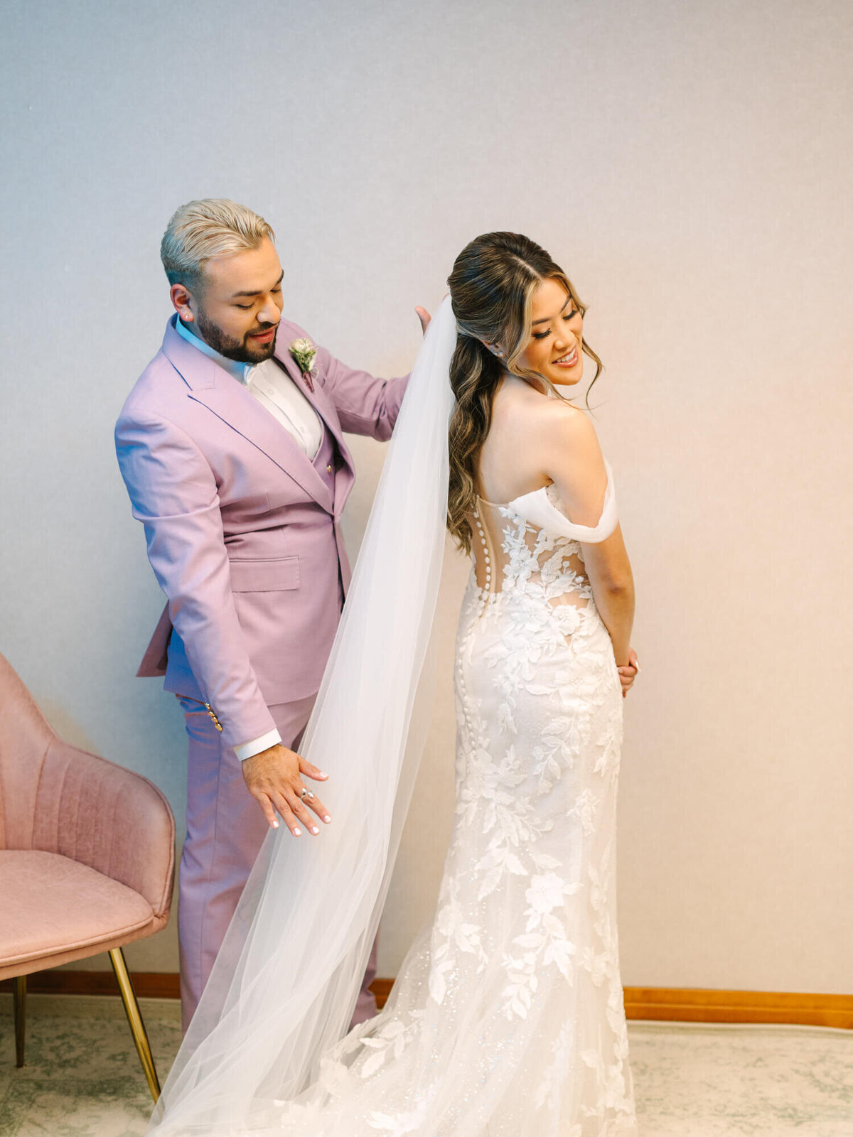 A bridesman in a lilac suit assists a smiling bride in an elegant, lace wedding dress. Her veil drapes gracefully, creating a joyful, intimate feel.