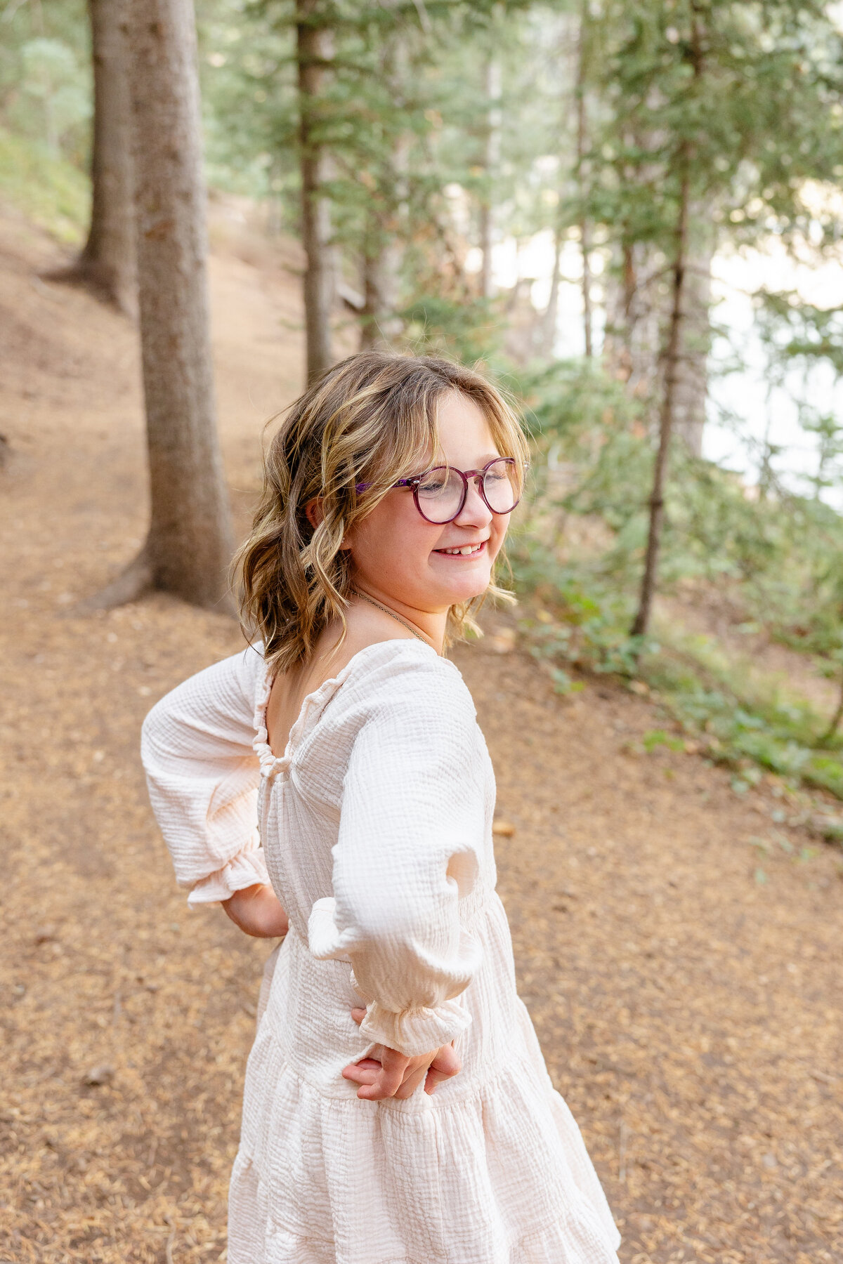 A young girl with glasses playfully poses with hands on her hips, wearing a light-colored dress. She stands in the forest, with soft natural light illuminating her.