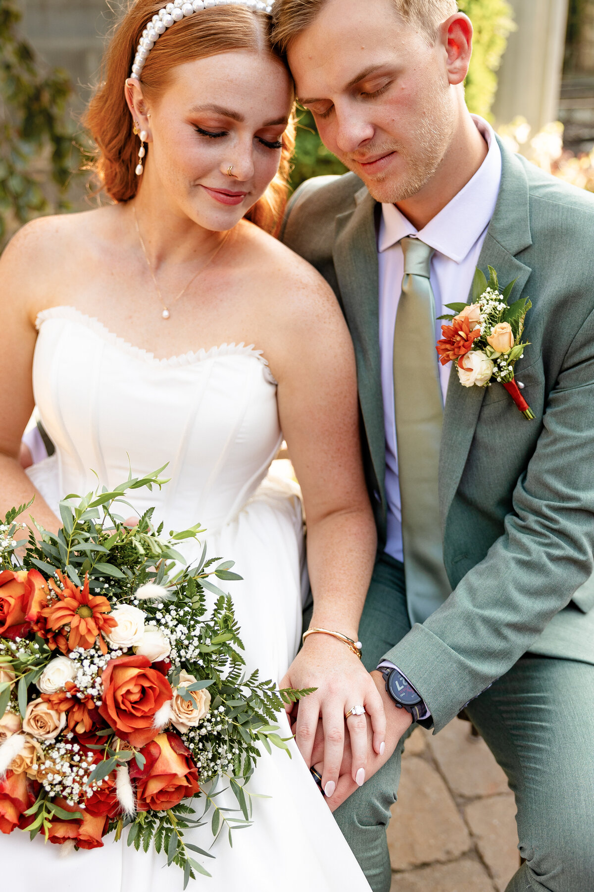 The bride and groom sit close together, admiring their wedding rings, with the bride’s vibrant orange and green bouquet resting in her lap.